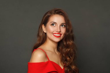 Gülümseyen esmer, modaya uygun makyajlı, kahverengi kıvırcık saçlı, dalgalı saçlı ve siyah arka planda kırmızı dudaklı bir kadın. 