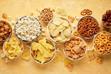 Parlak sarı arka planda farklı tuzlu atıştırmalıklar bulunan bir sürü kase. Cips, patlamış mısır, fındık, cips ve kraker. Parti yemeği. Üst görünüm