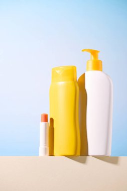 Açık mavi arka planda sarı ve beyaz güneş kremi şişeleri ve ruj. Güneş koruma ürünleri. Boş şişe maketi. Güneşli ışık.