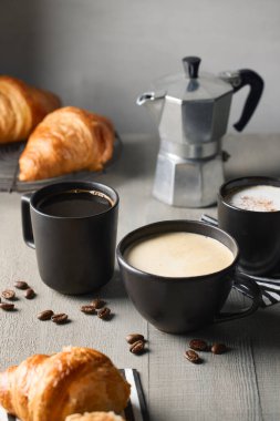 Gri ahşap masada üç fincan siyah kahve ve taze pişmiş kruvasan. Farklı bir kahve çeşidi. Americano, cappuccino ve latte.