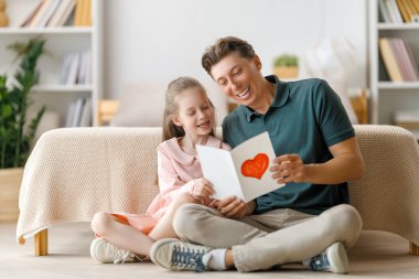 Çocuk kızı babasını tebrik ediyor ve ona kartpostal veriyor. Baba ve kız gülümsüyor ve sarılıyorlar. Aile tatili ve birliktelik.