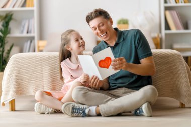 Çocuk kızı babasını tebrik ediyor ve ona kartpostal veriyor. Baba ve kız gülümsüyor ve sarılıyorlar. Aile tatili ve birliktelik.