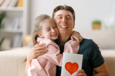 Çocuk kızı babasını tebrik ediyor ve ona kartpostal veriyor. Baba ve kız gülümsüyor ve sarılıyorlar. Aile tatili ve birliktelik.