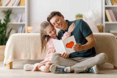 Çocuk kızı babasını tebrik ediyor ve ona kartpostal veriyor. Baba ve kız gülümsüyor ve sarılıyorlar. Aile tatili ve birliktelik.