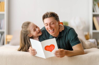 Çocuk kızı babasını tebrik ediyor ve ona kartpostal veriyor. Baba ve kız gülümsüyor ve sarılıyorlar. Aile tatili ve birliktelik.