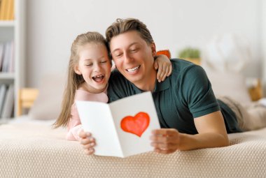 Çocuk kızı babasını tebrik ediyor ve ona kartpostal veriyor. Baba ve kız gülümsüyor ve sarılıyorlar. Aile tatili ve birliktelik.