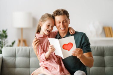 Çocuk kızı babasını tebrik ediyor ve ona kartpostal veriyor. Baba ve kız gülümsüyor ve sarılıyorlar. Aile tatili ve birliktelik.