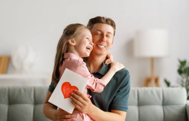 Çocuk kızı babasını tebrik ediyor ve ona kartpostal veriyor. Baba ve kız gülümsüyor ve sarılıyorlar. Aile tatili ve birliktelik.