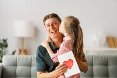 Çocuk kızı babasını tebrik ediyor ve ona kartpostal veriyor. Baba ve kız gülümsüyor ve sarılıyorlar. Aile tatili ve birliktelik.