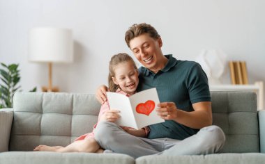 Çocuk kızı babasını tebrik ediyor ve ona kartpostal veriyor. Baba ve kız gülümsüyor ve sarılıyorlar. Aile tatili ve birliktelik.