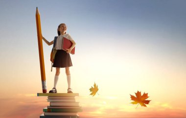 Okula dönün! Kitap kulesinde duran ve elinde günbatımı gökyüzünün arka planında kocaman bir kalem tutan mutlu, sevimli, çalışkan çocuk. Eğitim ve okuma kavramı. Hayal gücünün gelişimi..