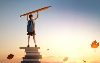 Okula dönün! Kitap kulesinde duran ve elinde günbatımı gökyüzünün arka planında kocaman bir kalem tutan mutlu, sevimli, çalışkan çocuk. Eğitim ve okuma kavramı. Hayal gücünün gelişimi..