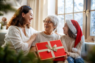 Mutlu Noeller ve mutlu tatiller. Neşeli çocuk, anneye ve büyükanneye hediyeler sunuyor. Ebeveynler ve küçük çocuklar kapalı alanda eğleniyorlar. Odasında hediyeler olan sevgi dolu bir aile.