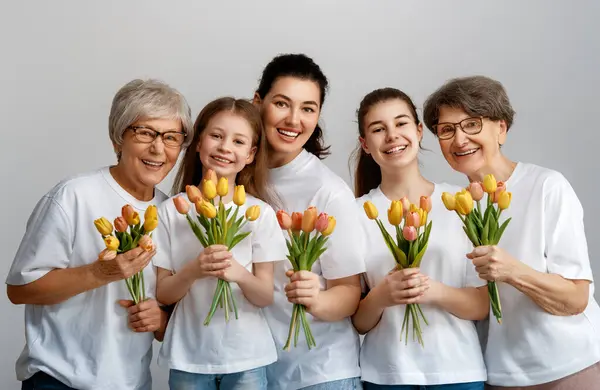 Happy women\'s day! Children daughters are congratulating mom and grandmothers giving them flowers tulips. Grannys, mom and girls smiling on light grey background. Family holiday and togetherness.