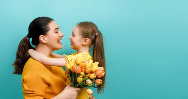 Happy women\'s day. Child is congratulating mom and giving her yellow flowers. Mum and girl smiling on teal background. Family holiday and togetherness.