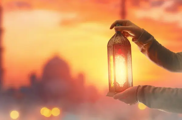 Woman is holding Ornamental Arabic lantern with burning candle glowing at night mosque background. Festive greeting card, invitation for Muslim holy month Ramadan Kareem.