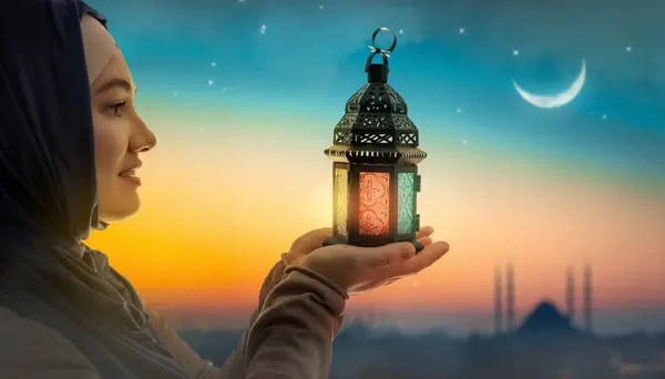 Woman is holding Ornamental Arabic lantern with burning candle glowing at night mosque background. Festive greeting card, invitation for Muslim holy month Ramadan Kareem.