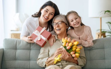 Anneler Günün kutlu olsun. Çocuk kızı ve annesi büyükannesinin ona çiçek lalesi ve hediye vermesini kutluyorlar. Büyükanne, anne ve kız gülümsüyor ve sarılıyorlar. Aile tatili ve birliktelik.