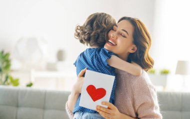 Anneler Günün kutlu olsun. Çocuk kızı annemi tebrik ediyor ve ona kartpostal veriyor. Anne ve kız gülümsüyor ve sarılıyorlar. Aile tatili ve birliktelik.