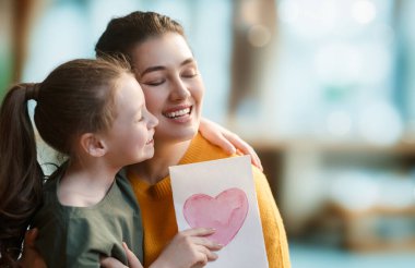 Anneler Günün kutlu olsun. Çocuk kızı annemi tebrik ediyor ve ona kartpostal veriyor. Anne ve kız gülümsüyor ve sarılıyorlar. Aile tatili ve birliktelik.