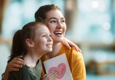 Anneler Günün kutlu olsun. Çocuk kızı annemi tebrik ediyor ve ona kartpostal veriyor. Anne ve kız gülümsüyor ve sarılıyorlar. Aile tatili ve birliktelik.