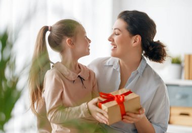 Anneler Günün kutlu olsun. Çocuk kızı annemi tebrik ediyor ve ona hediye paketi veriyor. Anne ve kız gülümsüyor ve sarılıyorlar. Aile tatili ve birliktelik.