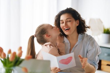 Anneler Günün kutlu olsun. Çocuk kızı annemi tebrik ediyor ve ona kartpostal veriyor. Anne ve kız gülümsüyor ve sarılıyorlar. Aile tatili ve birliktelik.