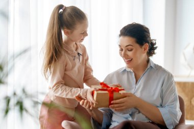 Anneler Günün kutlu olsun. Çocuk kızı annemi tebrik ediyor ve ona hediye paketi veriyor. Anne ve kız gülümsüyor ve sarılıyorlar. Aile tatili ve birliktelik.