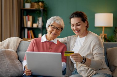 Genç ve yaşlı kadınlar online alışveriş için dizüstü bilgisayar kullanıyorlar. Kızı evde annesine yardım ediyor..