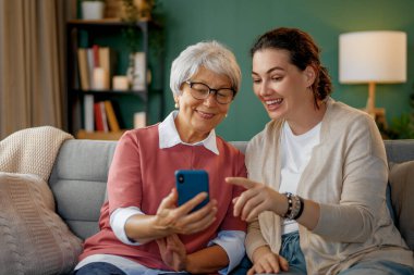 Genç ve yaşlı kadınlar akıllı telefon kullanıyor. Kız, anneye evde kalarak eğlenmesi için yardım ediyor..