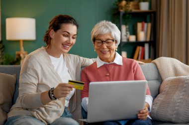 Genç ve yaşlı kadınlar online alışveriş için dizüstü bilgisayar kullanıyorlar. Kızı evde annesine yardım ediyor..