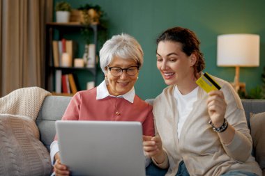 Genç ve yaşlı kadınlar online alışveriş için dizüstü bilgisayar kullanıyorlar. Kızı evde annesine yardım ediyor..