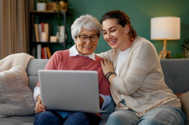 Genç ve yaşlı kadınlar dizüstü bilgisayar kullanıyorlar. Kız, anneye evde kalarak eğlenmesi için yardım ediyor..