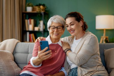 Genç ve yaşlı kadınlar akıllı telefon kullanıyor. Kız, anneye evde kalarak eğlenmesi için yardım ediyor..