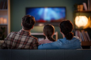 Happy family spending time together. Mother, father and child watching  TV, movies in the evening at home. clipart