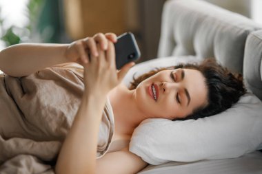 Mutlu, sıradan, güzel bir kadın evdeki yatakta telefon kullanıyor..