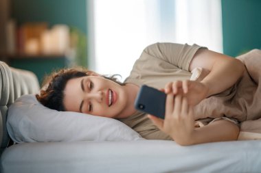 Mutlu, sıradan, güzel bir kadın evdeki yatakta telefon kullanıyor..