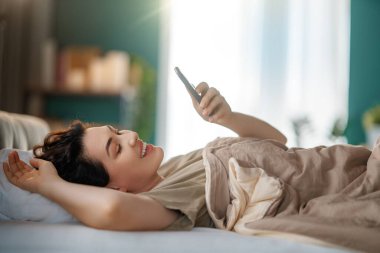 Mutlu, sıradan, güzel bir kadın evdeki yatakta telefon kullanıyor..