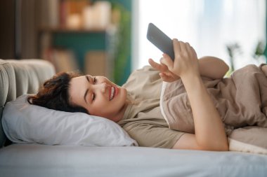 Mutlu, sıradan, güzel bir kadın evdeki yatakta telefon kullanıyor..