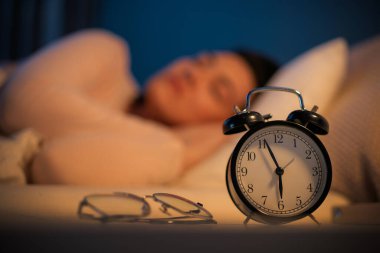 Kadın geceleri yatak odasında huzur içinde uyuyor. Ön planda alarm saati