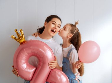Çocuk annesini tebrik ediyor ve ona pembe hava balonu veriyor. Anne ve kız açık gri arka planda gülümsüyor. Aile tatili ve birliktelik. Uluslararası Kadın Günü kavramı.