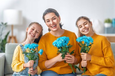 Anneler Günün kutlu olsun. Çocuklar annelerini tebrik ediyor ve ona çiçek veriyorlar. Anne ve kızlar gülümsüyor ve sarılıyorlar. Aile tatili ve birliktelik.