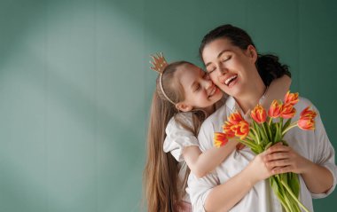Anneler Günün kutlu olsun! Çocuk kızı annemi tebrik ediyor ve ona çiçek laleleri veriyor. Anne ve kız yeşil arka planda gülümsüyor. Aile tatili ve birlikte