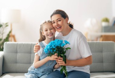 Anneler Günün kutlu olsun. Çocuk kızı annemi tebrik ediyor ve ona çiçek veriyor. Anne ve kız gülümsüyor ve sarılıyorlar. Aile tatili ve birliktelik.