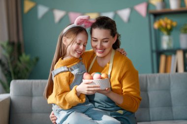 İyi tatiller! Anne ve kızı yumurta boyuyor. Paskalyayı kutlayan aile. Sevimli küçük kız tavşan kulağı takıyor..