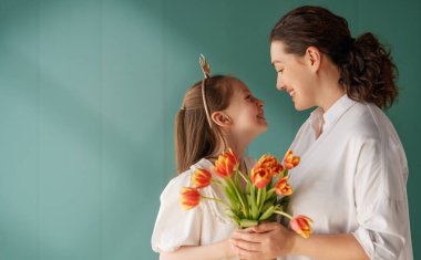 Anneler Günün kutlu olsun! Çocuk kızı annemi tebrik ediyor ve ona çiçek laleleri veriyor. Anne ve kız yeşil arka planda gülümsüyor. Aile tatili ve birlikte