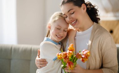 Anneler Günün kutlu olsun. Çocuk kızı annemi tebrik ediyor ve ona çiçek veriyor. Anne ve kız gülümsüyor ve sarılıyorlar. Aile tatili ve birliktelik.