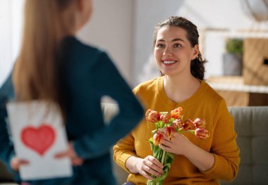 Anneler Günün kutlu olsun. Çocuk kızı annemi tebrik ediyor ve ona çiçek ve kartpostal veriyor. Anne ve kız gülümsüyor. Aile tatili ve birliktelik.