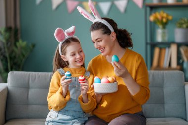 İyi tatiller! Anne ve kızı yumurta boyuyor. Paskalyayı kutlayan aile. Sevimli küçük kız tavşan kulağı takıyor..