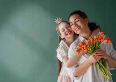 Anneler Günün kutlu olsun! Çocuk kızı annemi tebrik ediyor ve ona çiçek laleleri veriyor. Anne ve kız yeşil arka planda gülümsüyor. Aile tatili ve birlikte